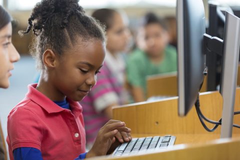 Elementary age African American little girl is smiling while typing on keyboard in public school library or computer lab. Student is using a desktop computer with friend to work on school assignment. She is wearing casual clothing.